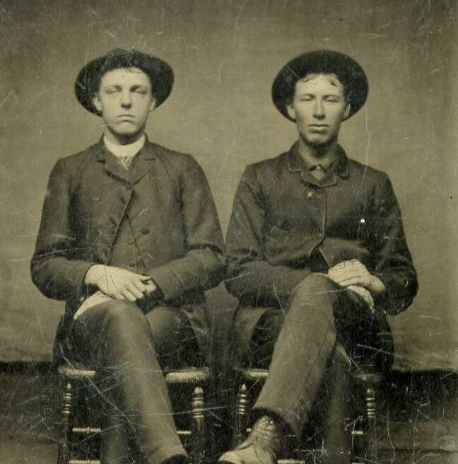 Black and white tintype photograph of two seated men in nearly identical outfits that include wide brimmed hats.